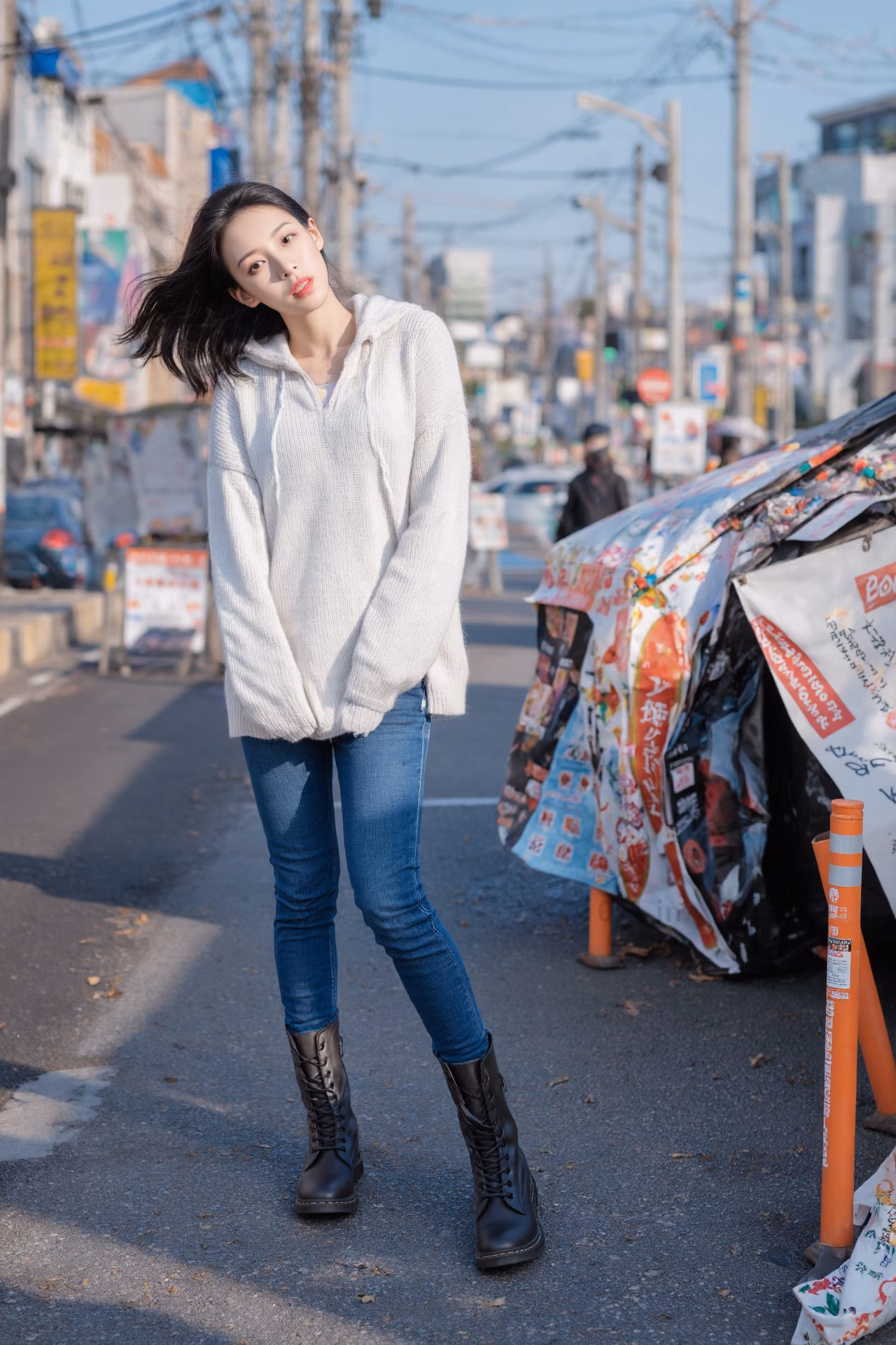 Close-up portrait of a slim Japanese woman wearing an oversized hoodie and bootcut jeans, surrounded by a Japanese street backdrop.